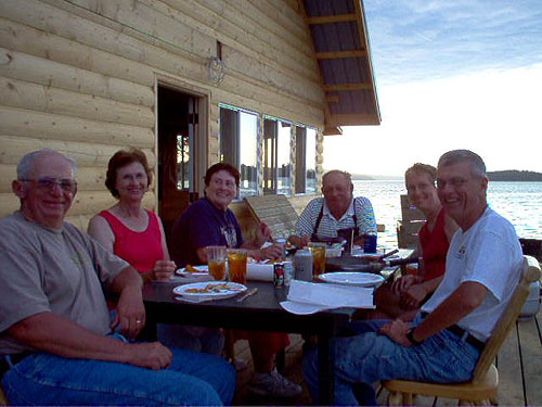 This is a photo of a group of people eating dinner on a porch right on the water and the sun is setting
